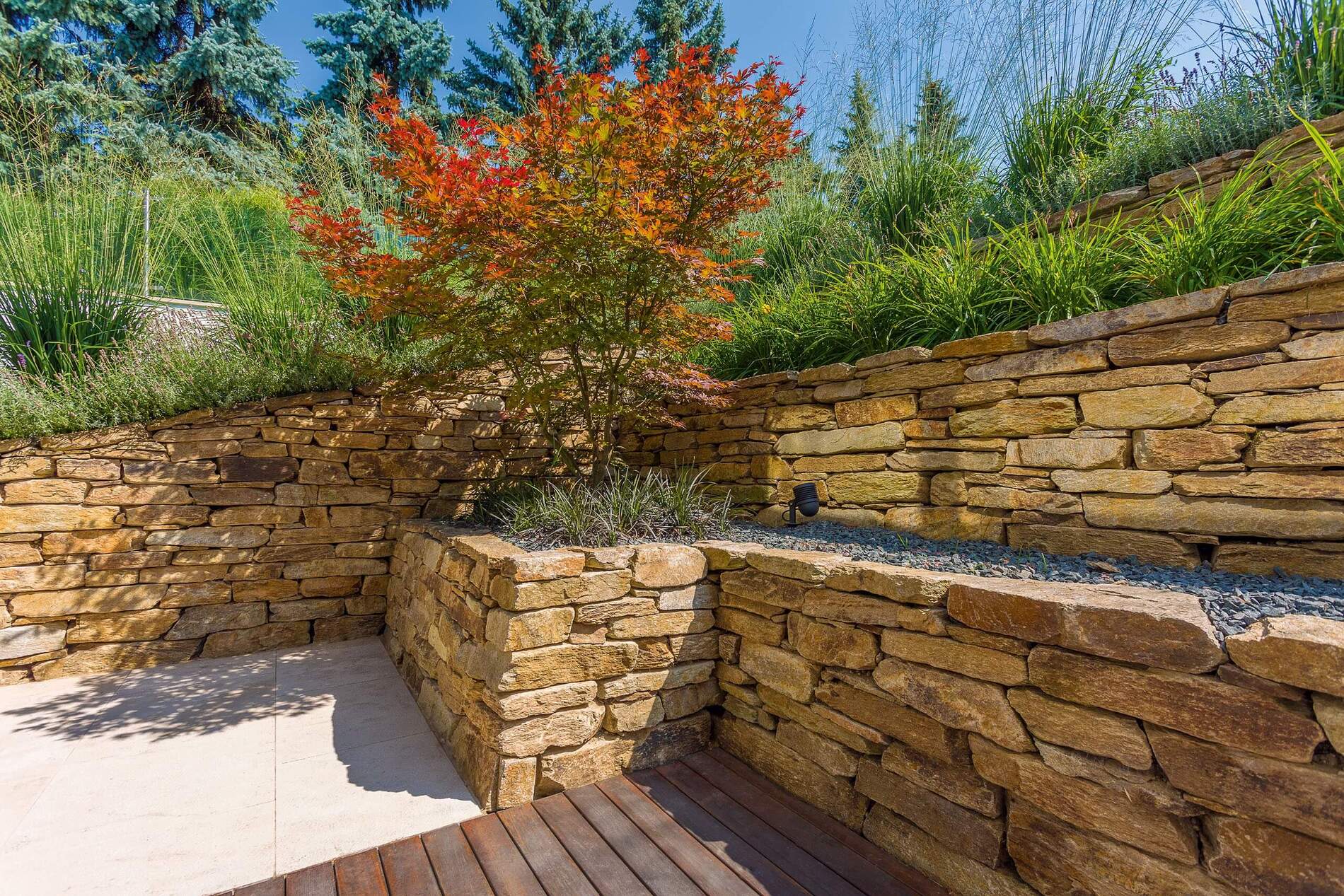 Gestaltung mit Natursteinmauer, Pflanzen und Holzbelag im Garten