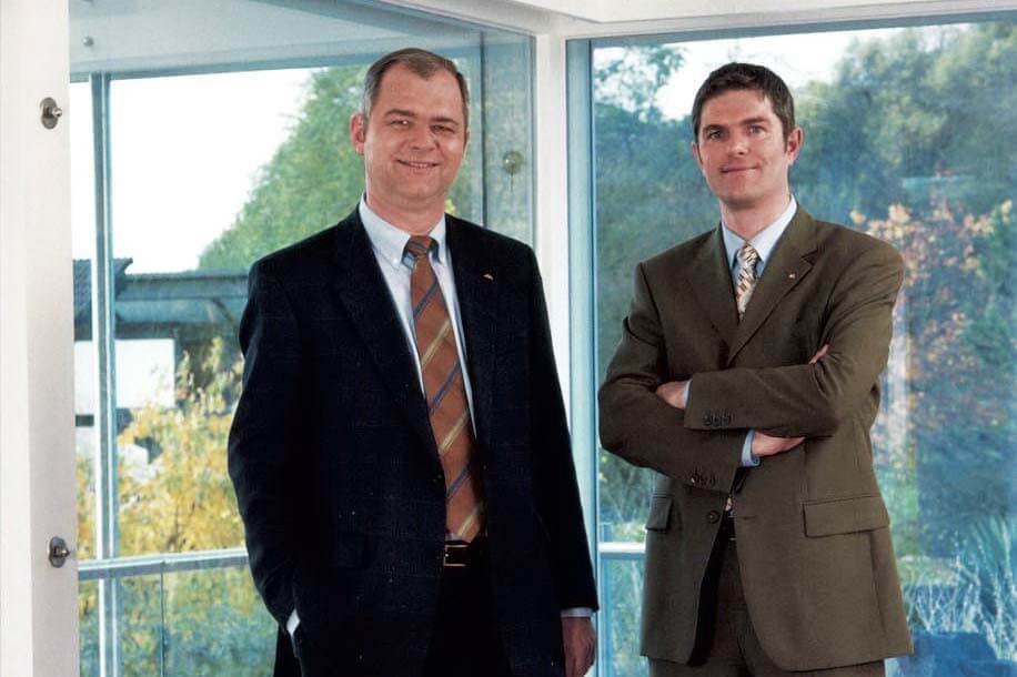 Zwei Männer in formeller Kleidung in einem modernen Gebäude Porträt zweier Männer, einer lächelt, vor Fenster mit Landschaftsblick