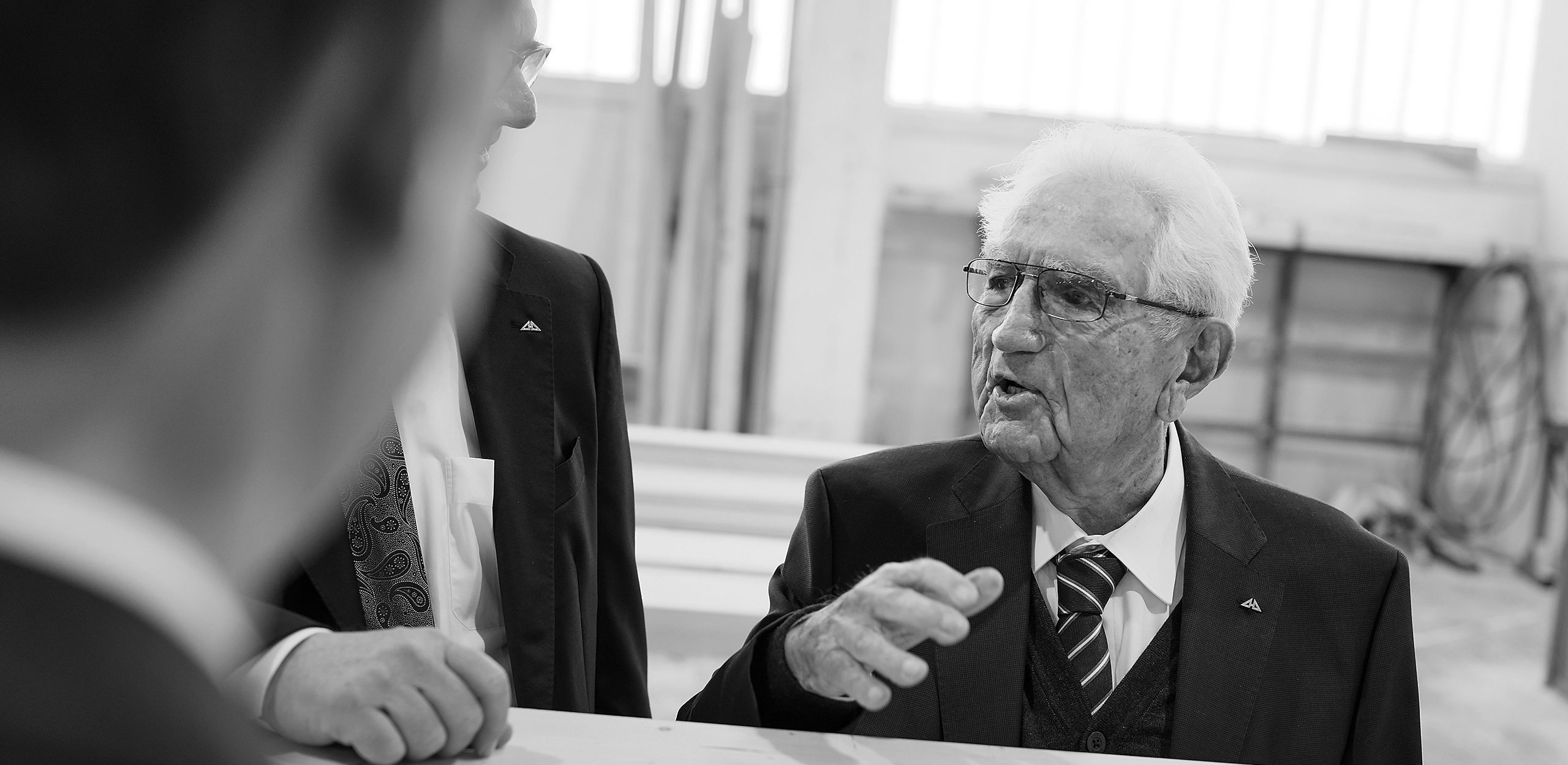 Älterer Mann spricht in einer professionellen Umgebung Ein älterer Mann erklärt etwas in einer Besprechung, im Hintergrund andere Personen.