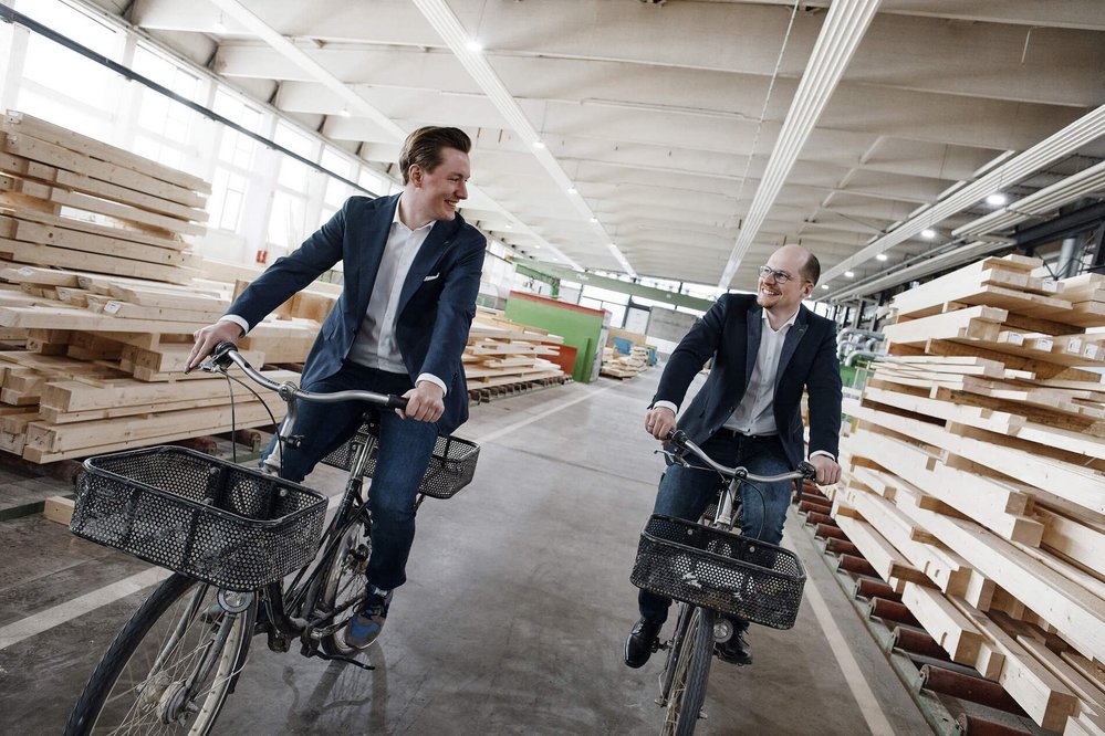 Zwei Männer auf Fahrrädern in einer Holzlagerhalle Zwei Männer lächeln, während sie in einer Holzlagerhalle Fahrrad fahren.