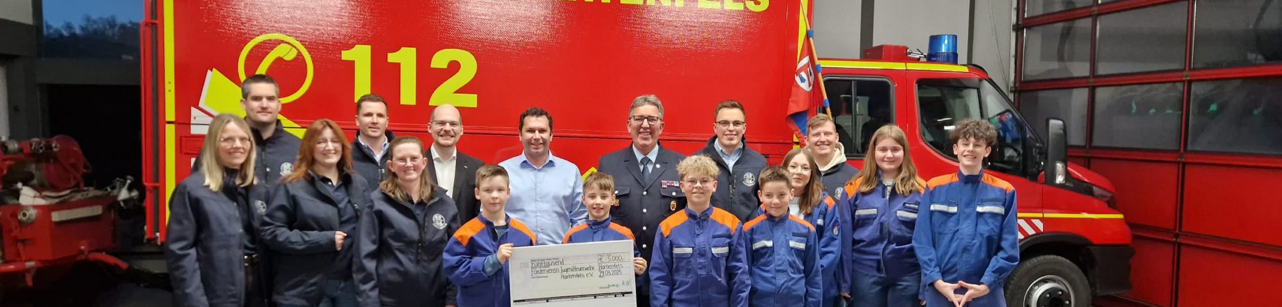 Gruppenfoto mit Feuerwehrteam und Scheck vor rotem Fahrzeug