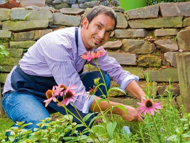 Mann im Garten mit Blumen, Holz und Steinen im Hintergrund.