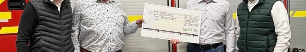 Vier Personen stehen vor einem Feuerwehrauto und halten einen Scheck.