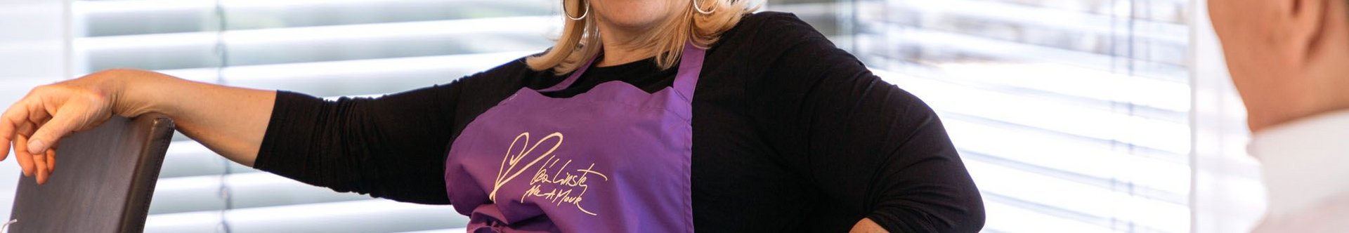 Frau in lila Schürze sitzt an einem Tisch und lächelt.