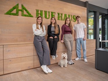Teamfoto im Büro von HUF HAUS mit Hund Vier Personen posieren vor einem modernen Büro mit Pflanzenlogo