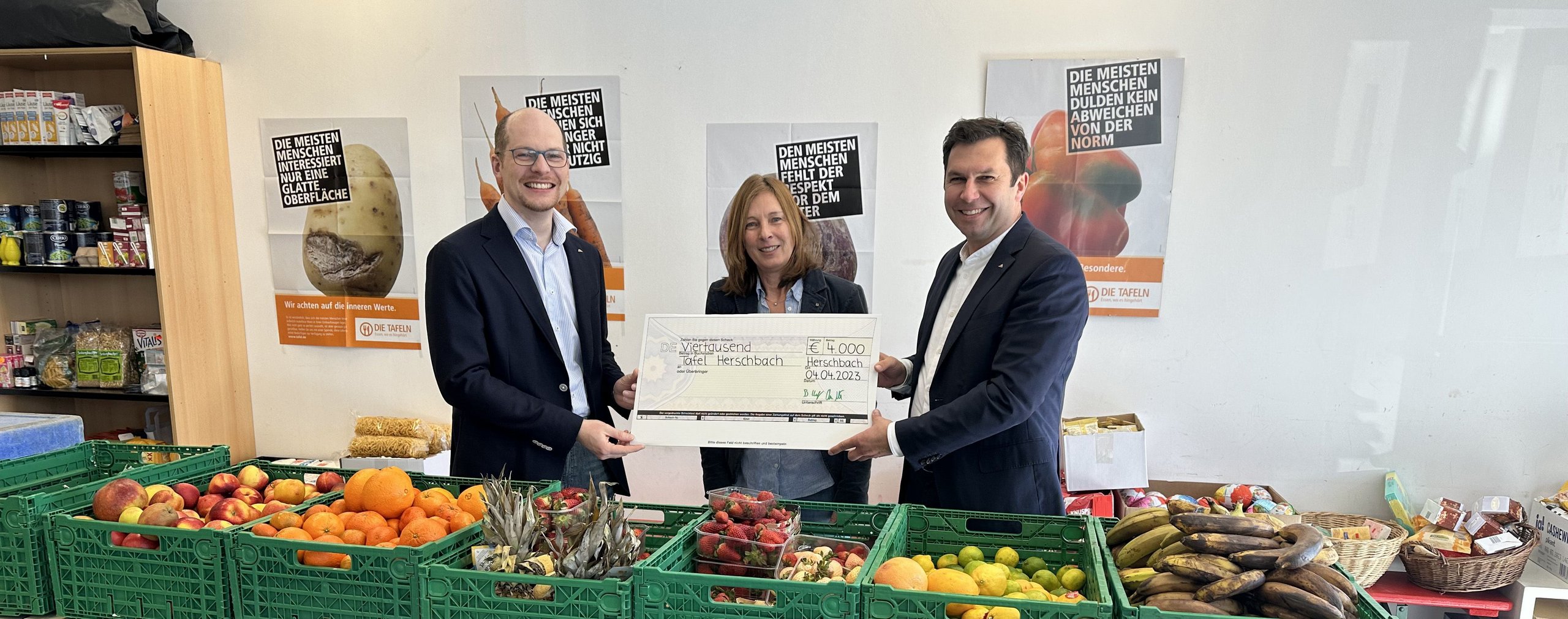 Drei Personen übergeben einen großen Scheck vor Obst und Gemüse