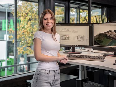 Junge Frau am Arbeitsplatz mit Computerbildschirmen Junge Frau posiert in modernem Büro mit Architekturvisualisierungen