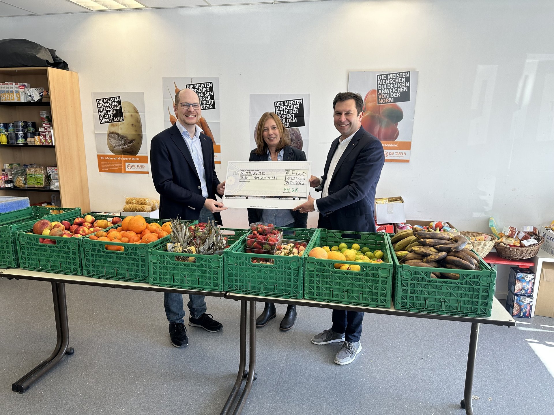 Drei Personen übergeben einen großen Scheck in einem Raum mit Obst und Gemüse