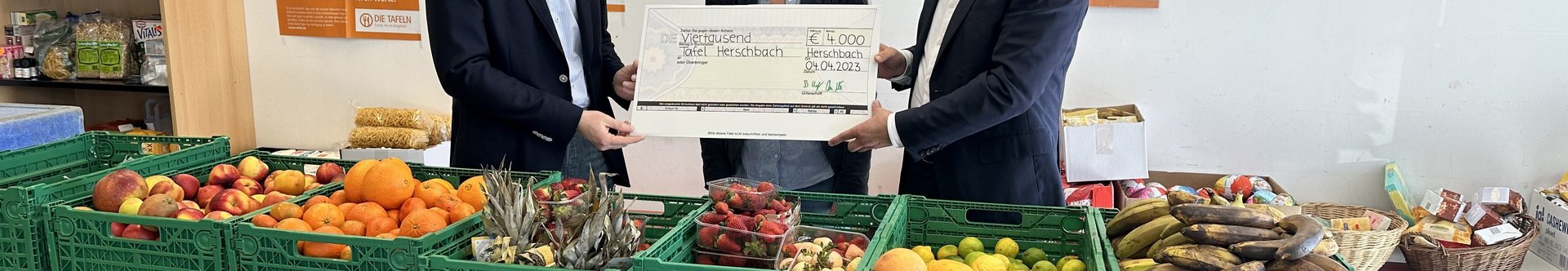 Drei Personen übergeben einen großen Scheck in einem Raum mit Obst und Gemüse