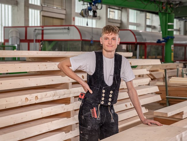 Junger Holzarbeiter in einer Werkstatt Ein junger Mann steht neben gestapeltem Holz in einer Werkstatt.